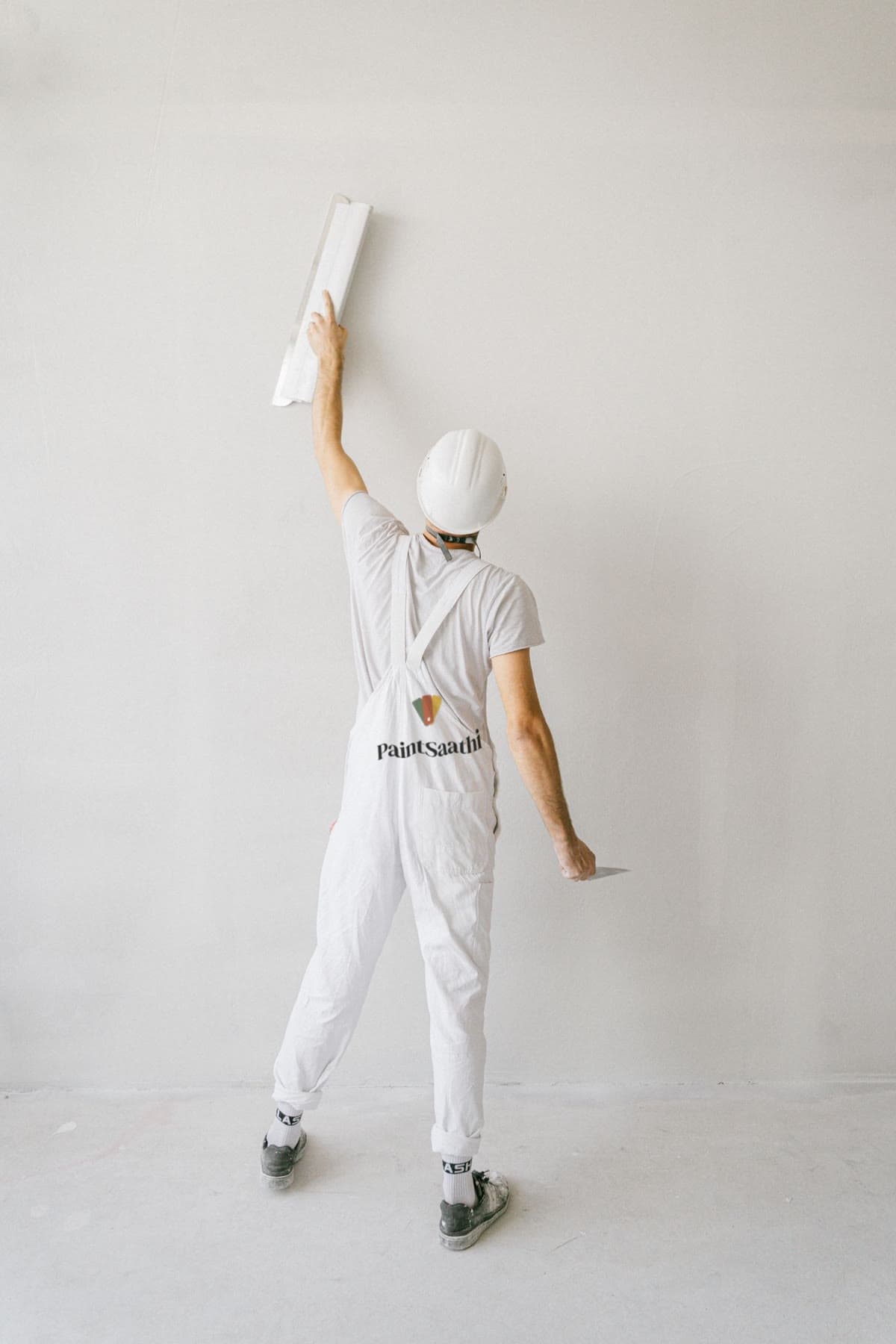 Painter in a white Paint Saathi uniform, seen from behind, smoothing a wall with a long trowel