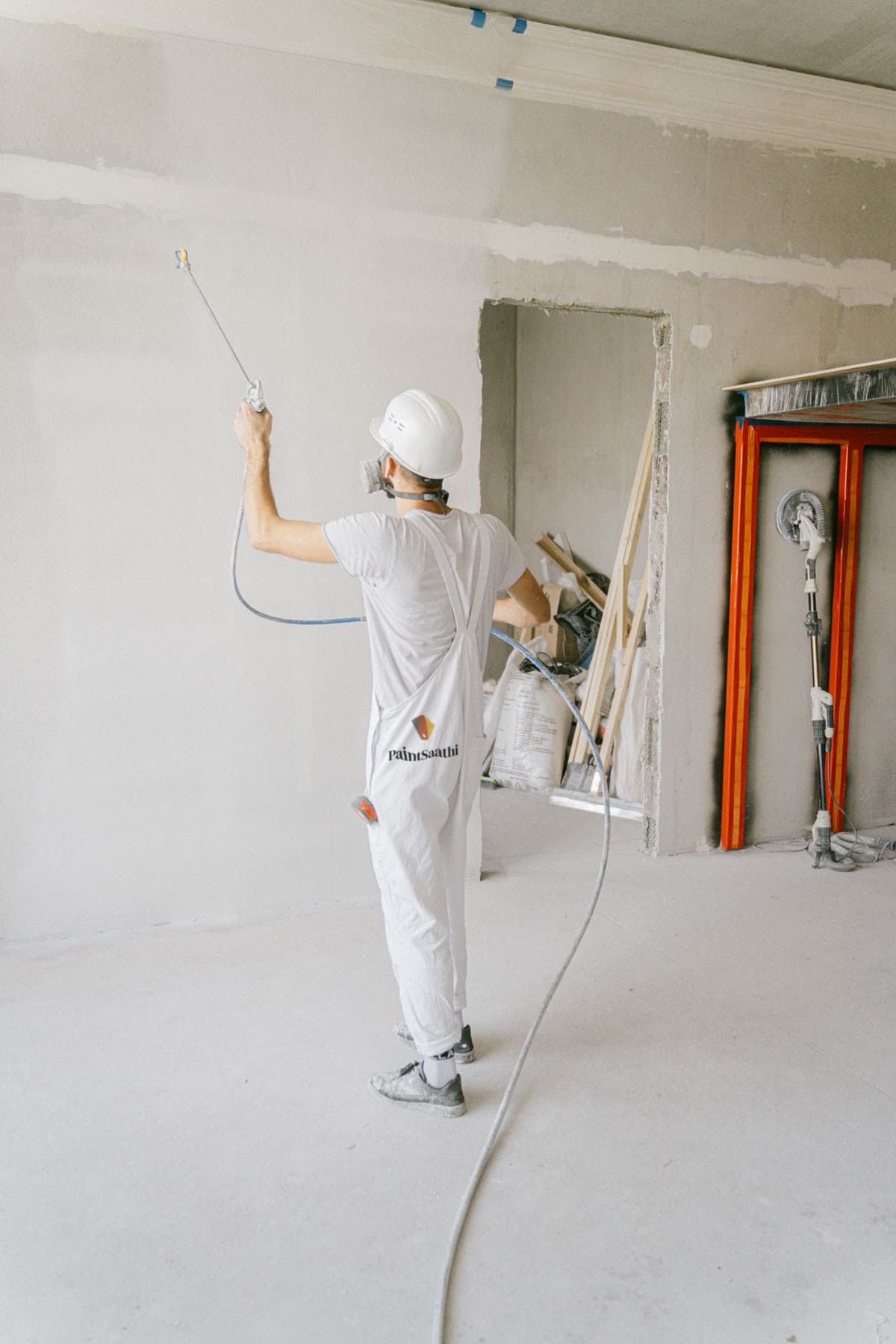 Painter in a white Paint Saathi uniform spraying primer onto a bare wall in an unfinished room