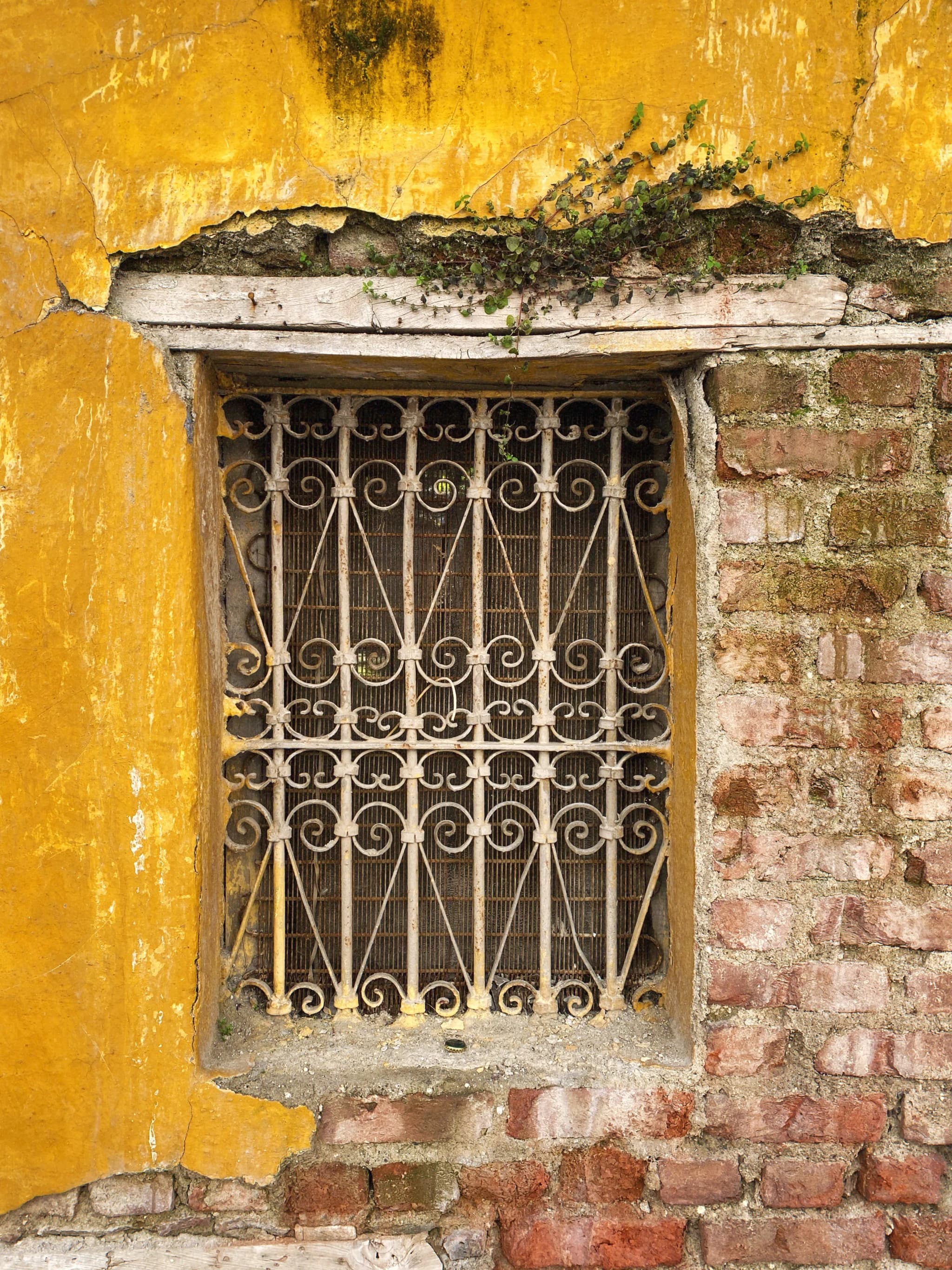 Weathered yellow wall with flaking plaster and exposed brick around an iron window grill