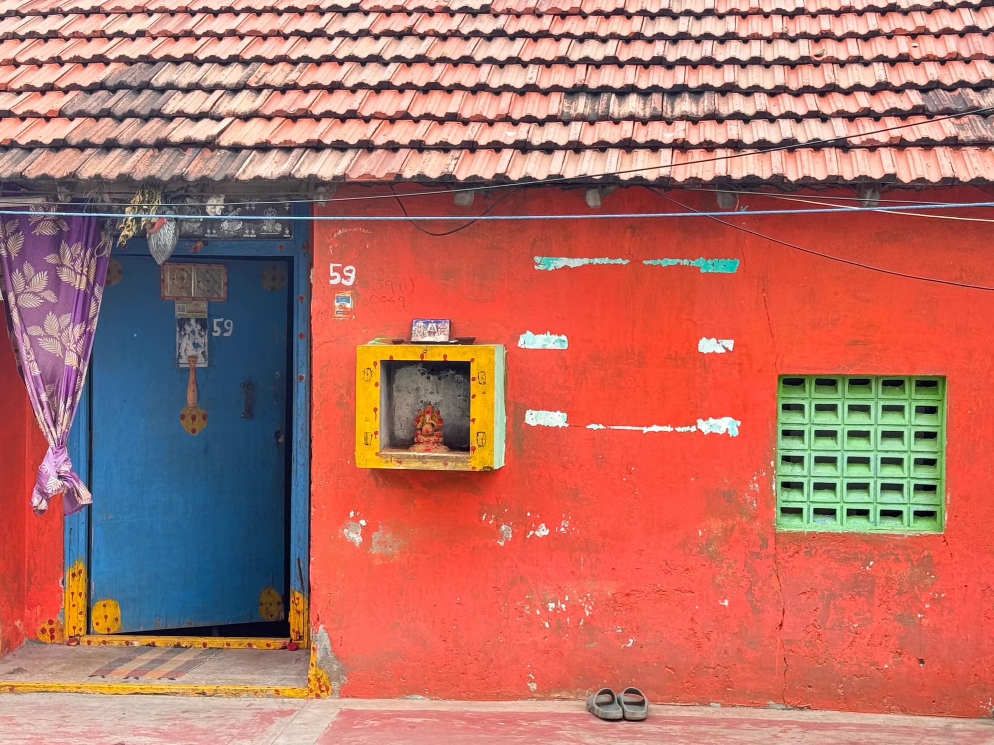Red house wall with a blue door, a small yellow shrine and a green jali window under a tiled roof