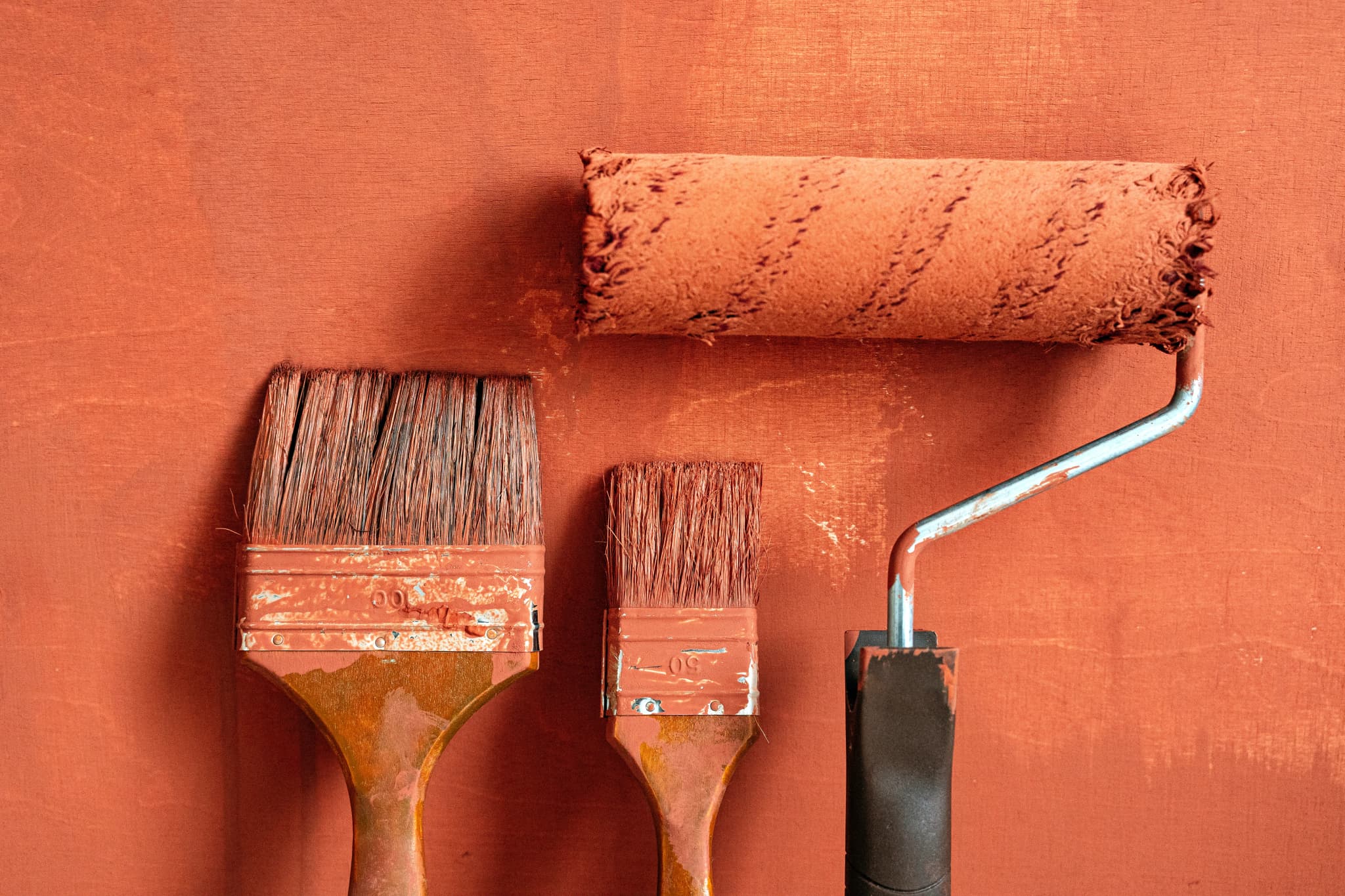 Two brushes and a roller loaded with terracotta paint, propped against a wall of the same colour