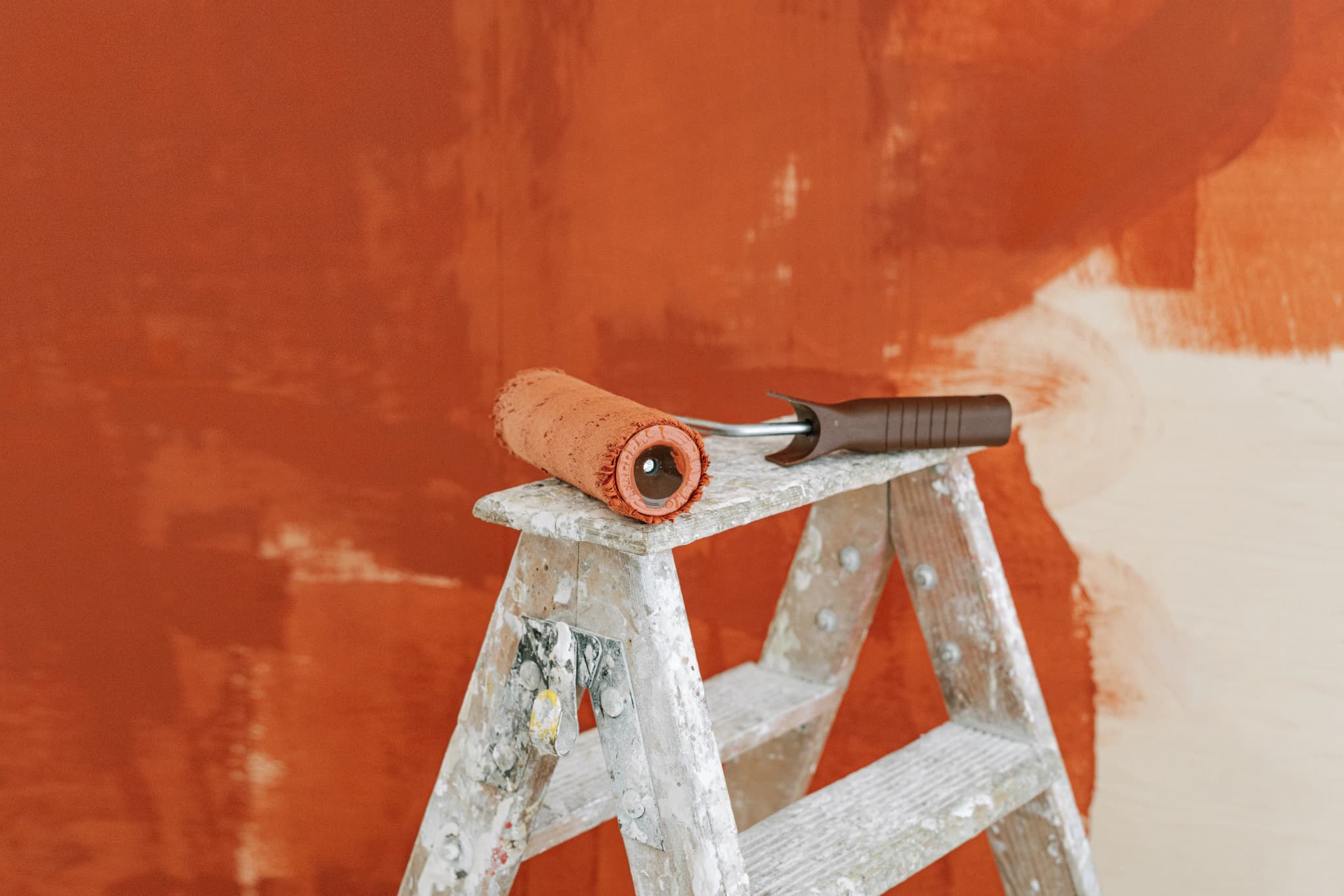 Paint roller resting on a stepladder in front of a wall half covered in terracotta paint
