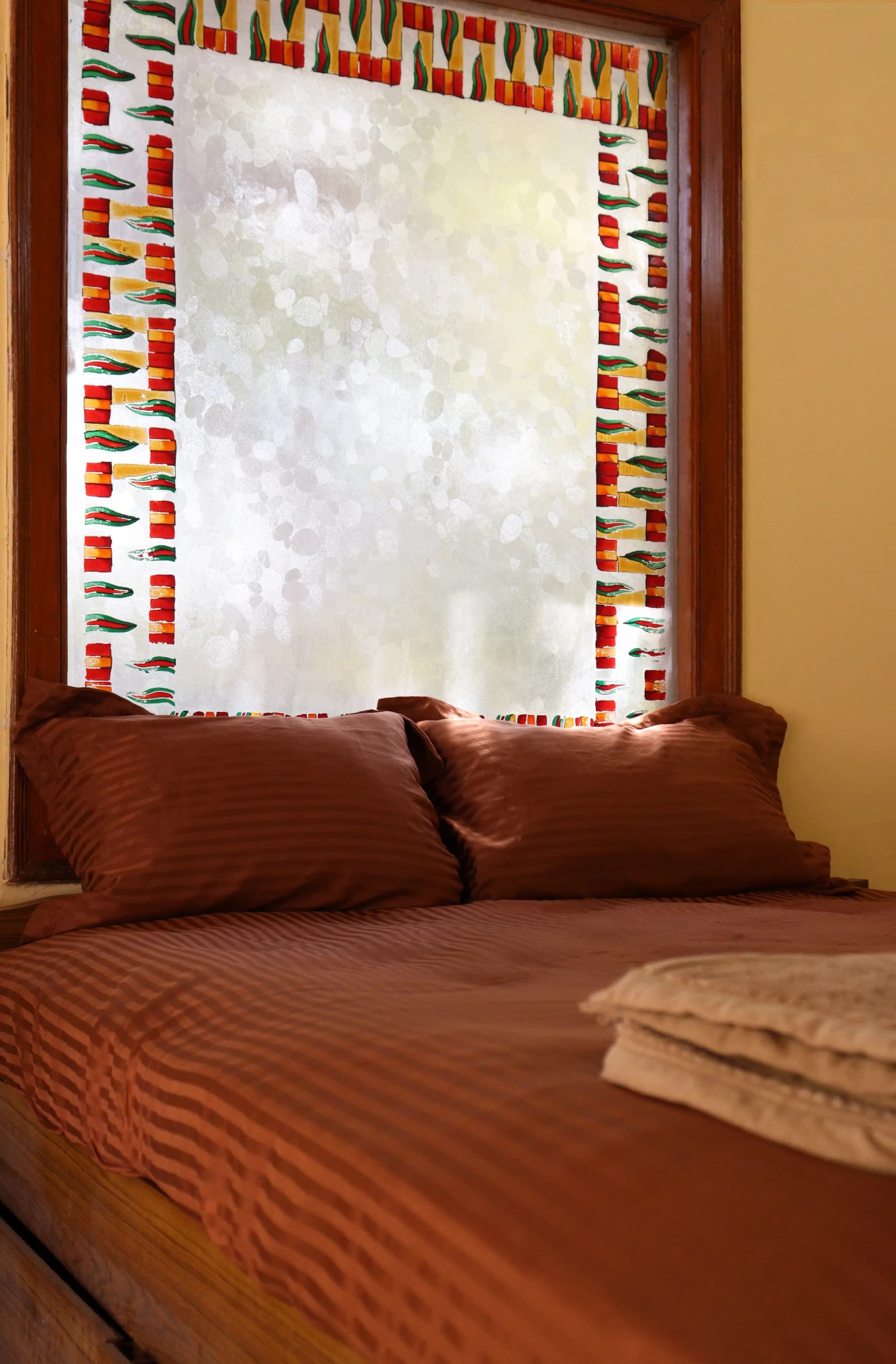 Bedroom with warm ochre walls, rust-brown bedding and a window framed in coloured glass